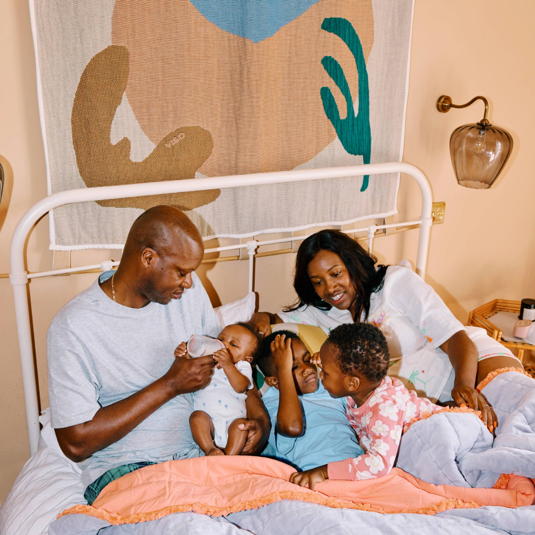 Family sat in bed mum is using a handsfree breast pump to express milk and store it in the fridge, dad is feeding baby expressed breast milk from a Pippeta breast milk storage bottle with teat